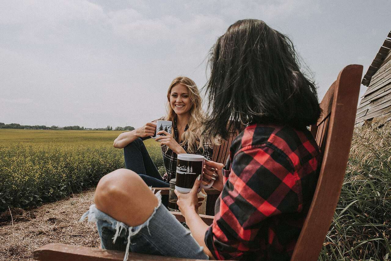 Zwei junge Frauen sitzen auf Stühlen in einem Feld und trinken Kaffee aus Bechern.