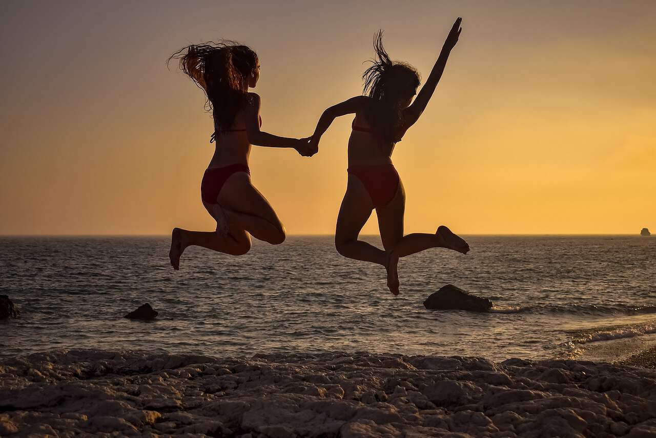 Zwei junge Frauen springen bei Sonnenuntergang am Strand vor Freude in die Luft.
