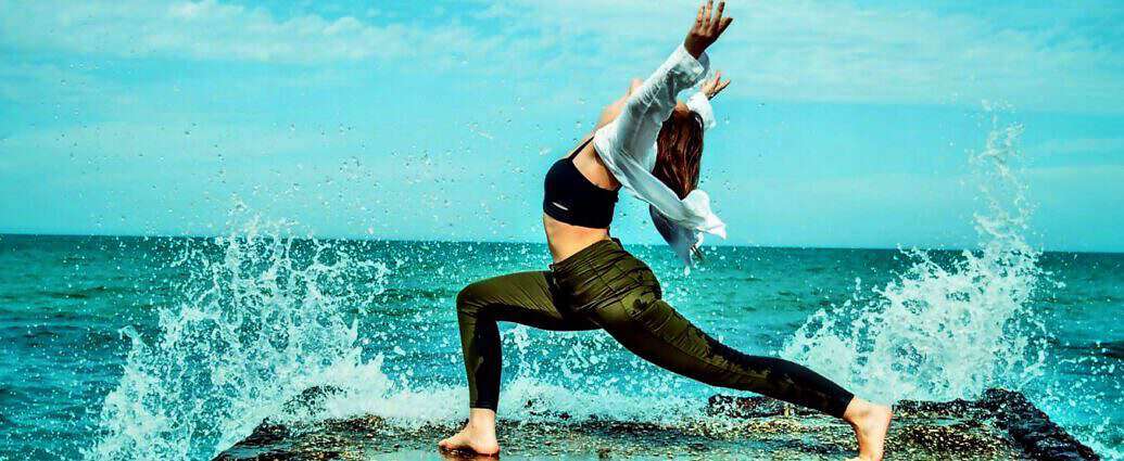 Eine junge Frau in grünen Jeans verrenkt ihren Körper in einer Pose der Freude direkt am Meer.