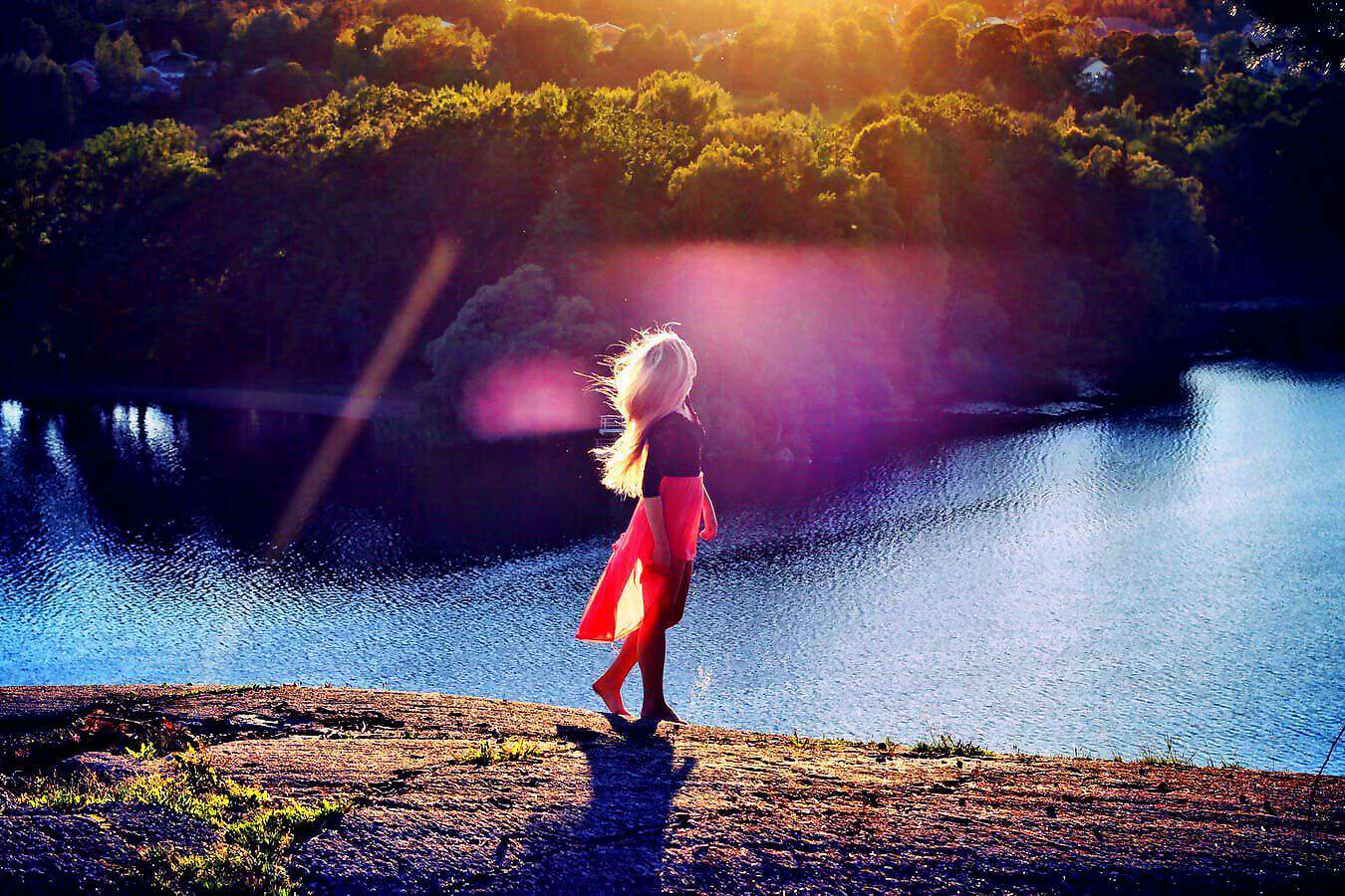 Eine blonde junge Frau geht nachdenklich an einem See spazieren.