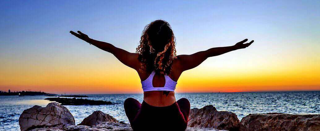 Eine junge Frau sitzt mit ausgestreckten Armen am Meer.