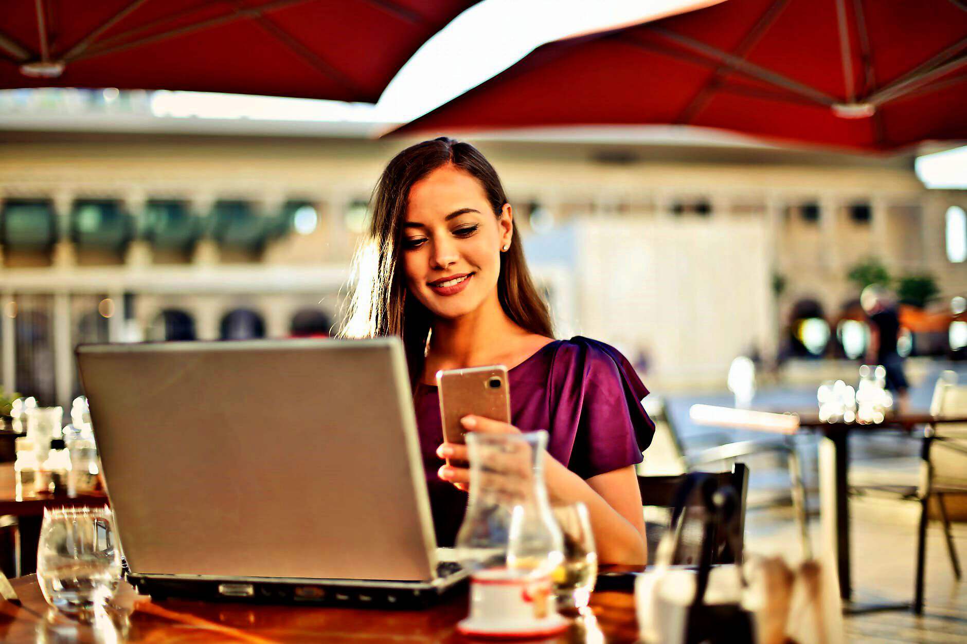 Eine junge Frau sitzt am Schreibtisch vor ihrem Laptop und schaut auf ihr Smartphone.
