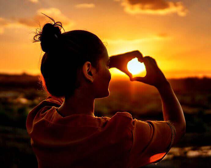 Eine junge Frau formt ihre Hände zu einem Herzen und schaut durch einen Sonnenuntergang.