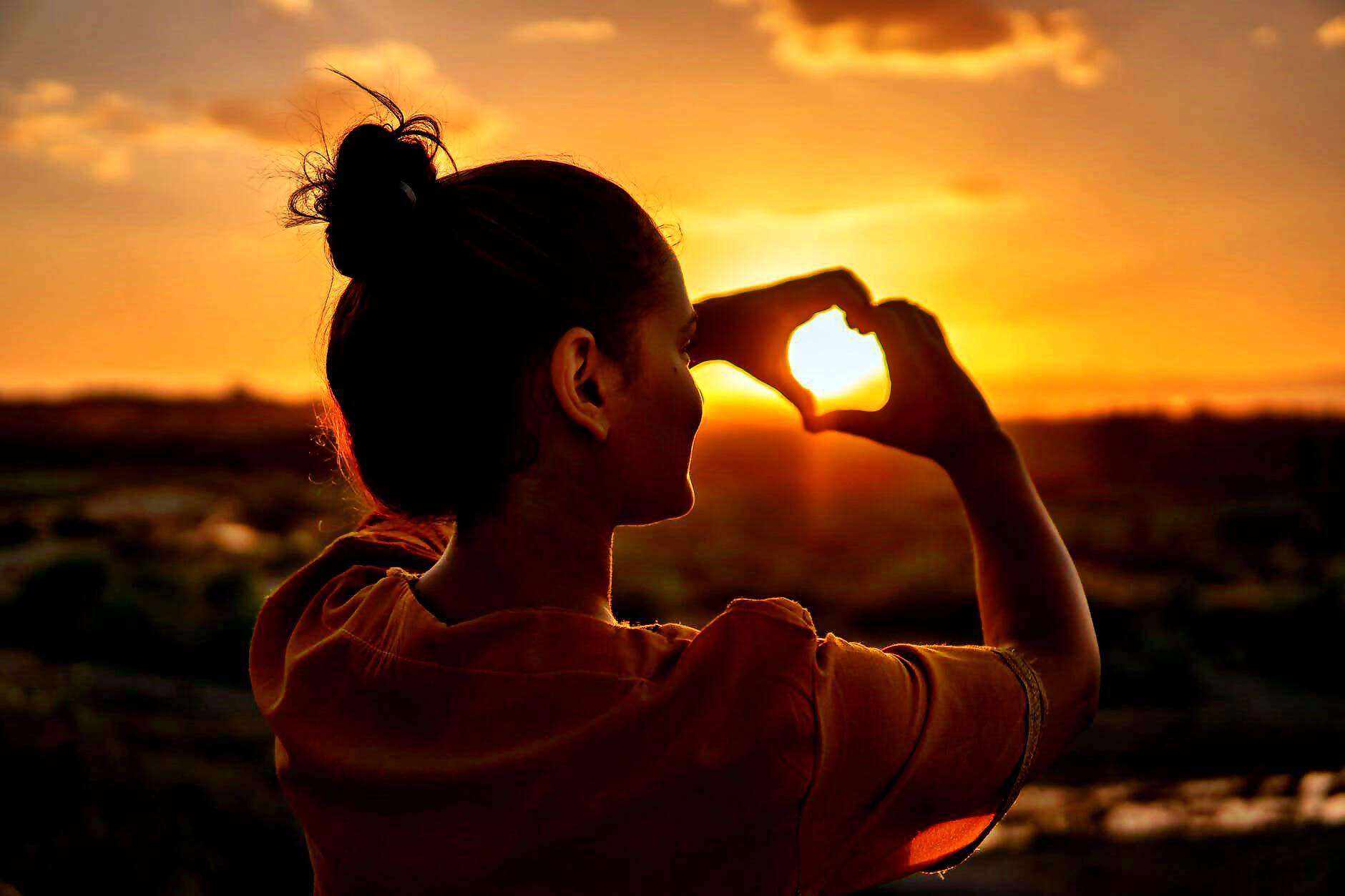 Eine junge Frau formt ihre Hände zu einem Herzen und schaut durch einen Sonnenuntergang.
