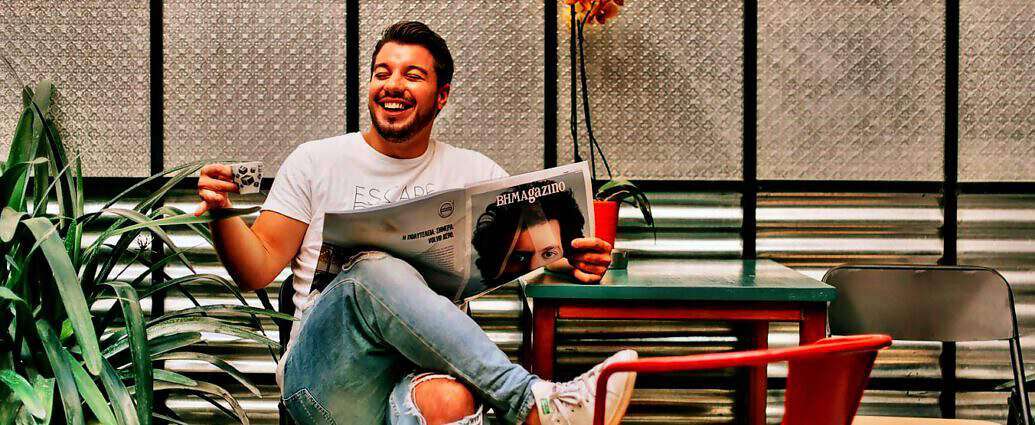 Ein junger Italiener mit einer Zeitschrift und einer Tasse Espresso in der Hand lacht fröhlich.