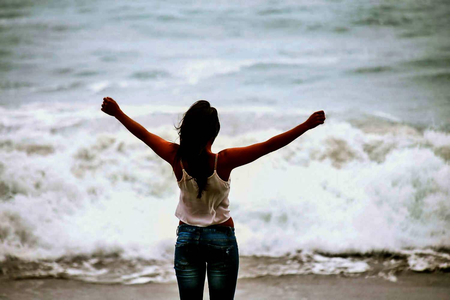 Eine junge Frau steht am Meer und streckt vor Freude ihre Arme aus.