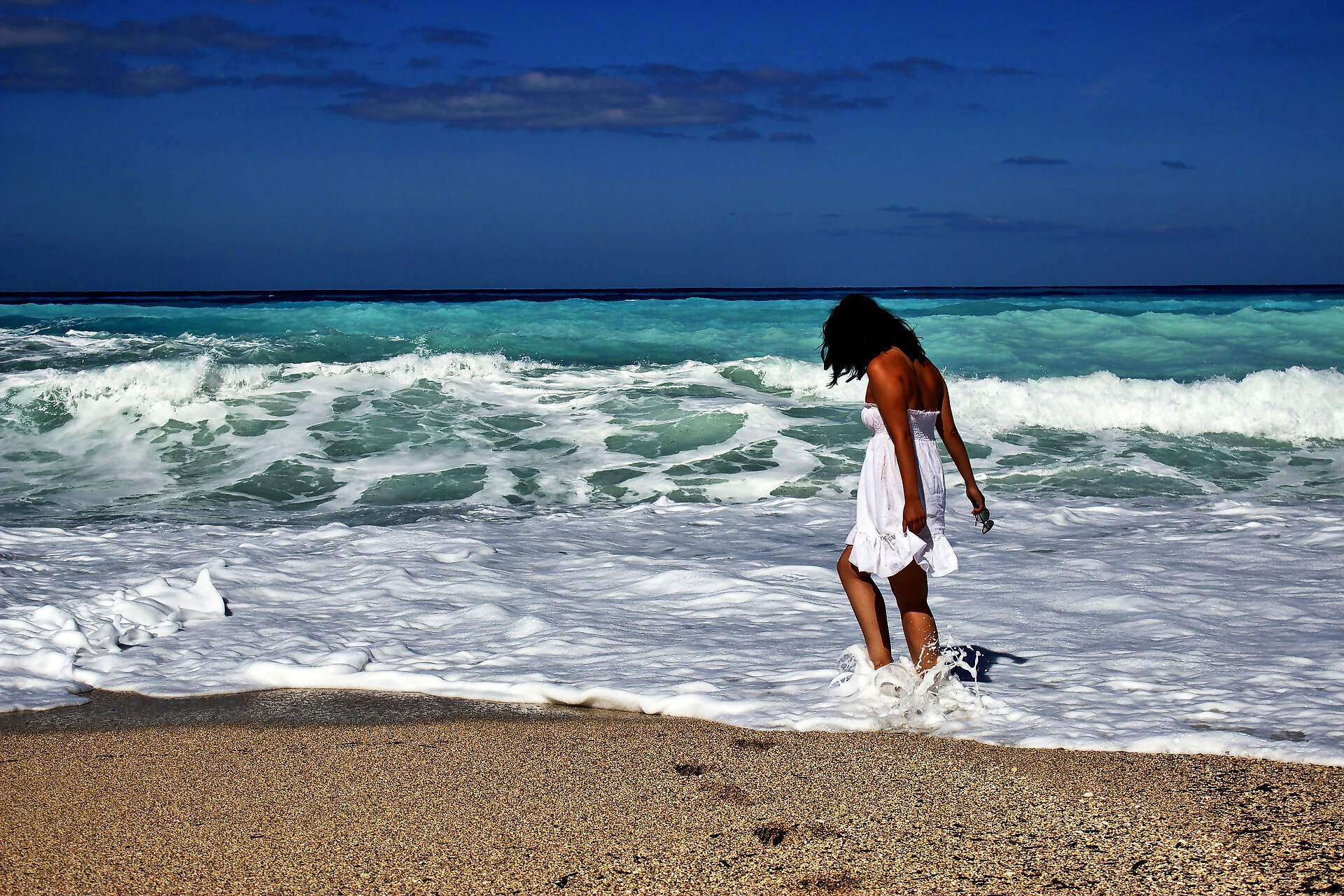 Eine junge Frau in weißem Kleid spielt am Strand mit ihren Füßen im Wasser.