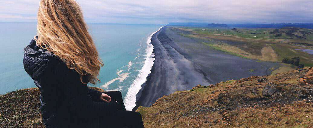 Eine junge Frau sitzt auf einer Düne und blickt von oben auf den Strand und das Meer.