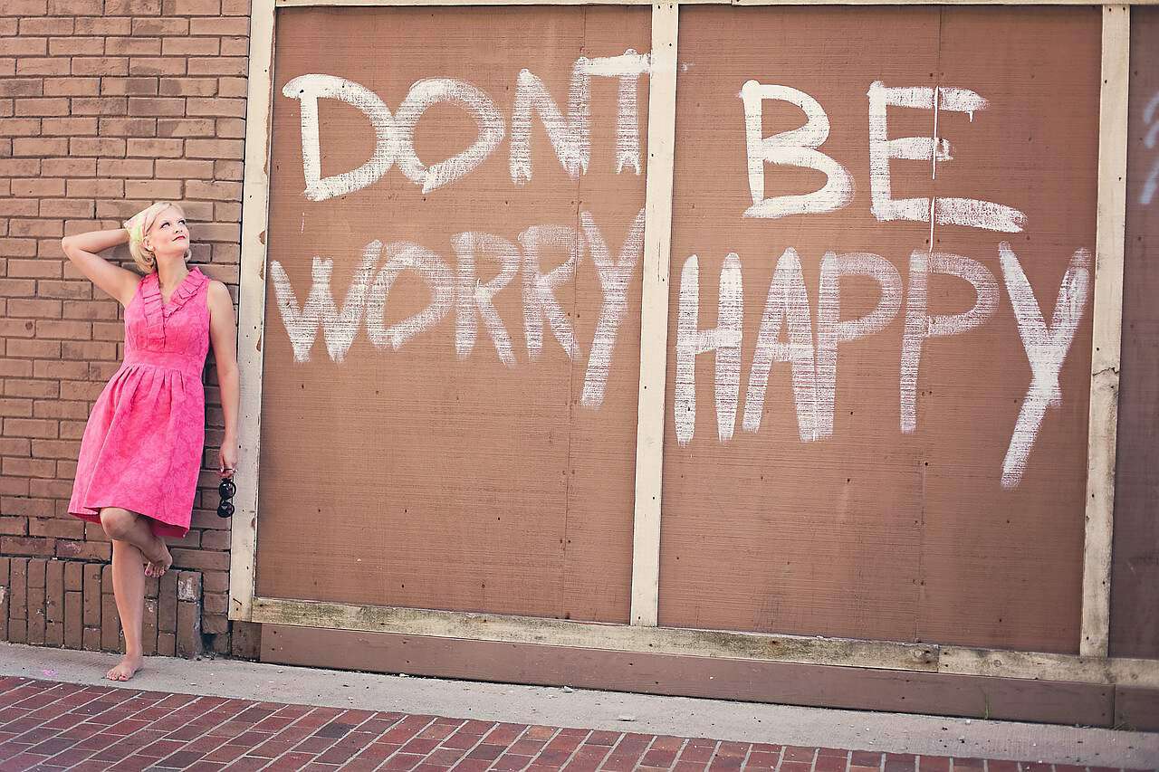 Eine junge blonde Frau in einem rosa Kleid steht an einer Wand mit der Aufschrift Dont Worry Be Happy.