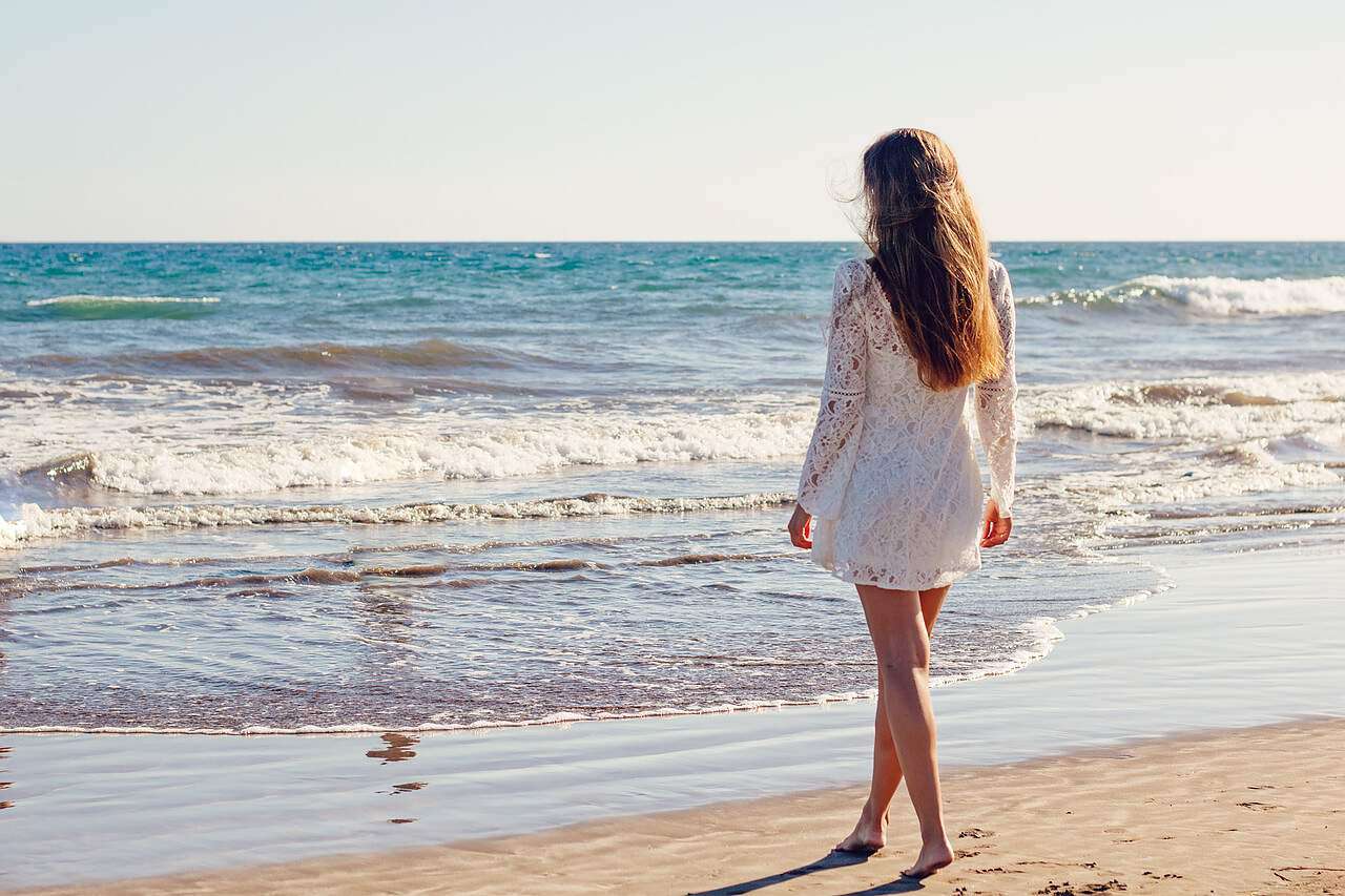 Am Strand steht eine Frau im weißen Strandkleid und schaut auf das Meer.