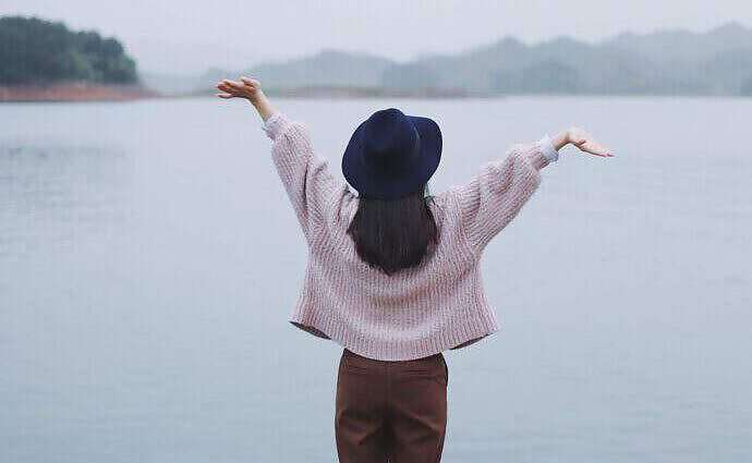Eine junge Frau steht vor einem See und hebt vor Freude die Arme hoch.