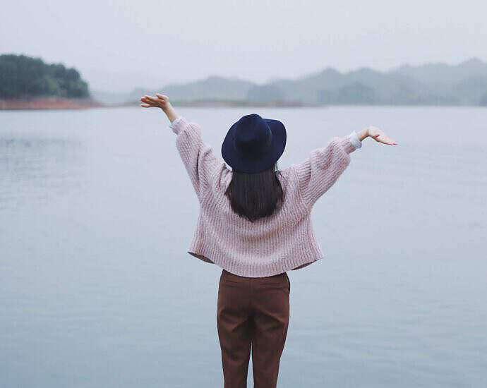 Eine junge Frau steht vor einem See und hebt vor Freude die Arme hoch.