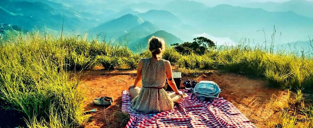 Eine schöne, junge Frau sitzt mit einem Kleid auf einer Wolldecke und picknickt, während sie auf die Berge schaut.