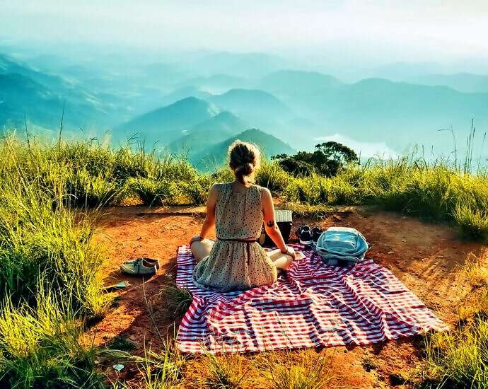 Eine schöne, junge Frau sitzt mit einem Kleid auf einer Wolldecke und picknickt, während sie auf die Berge schaut.