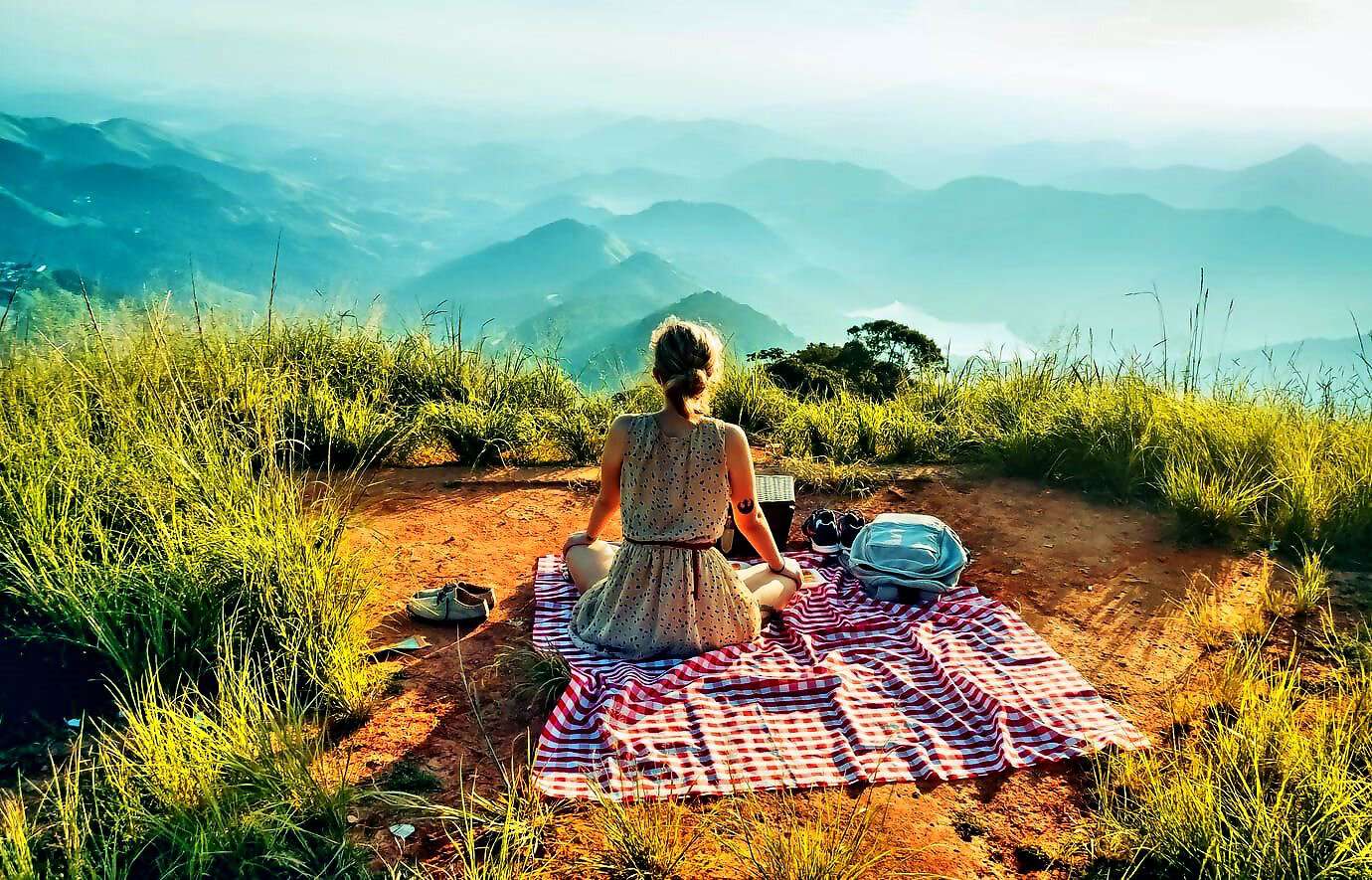Eine schöne, junge Frau sitzt mit einem Kleid auf einer Wolldecke und picknickt, während sie auf die Berge schaut.