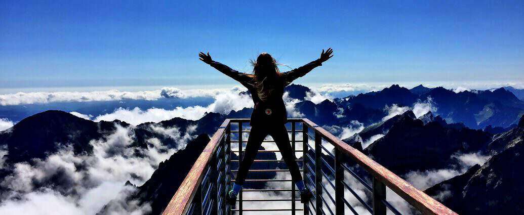 Eine junge Frau steht auf einer Plattform. Unter ihr erstrecken sich die Berge.