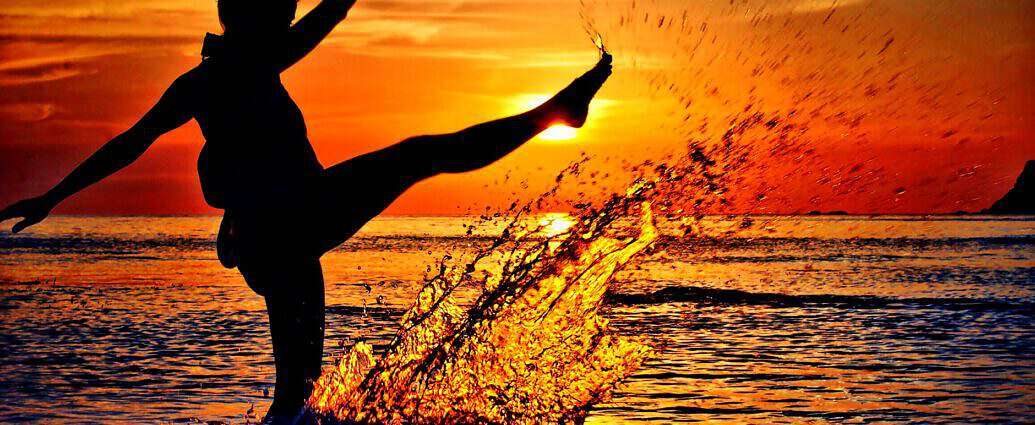 Eine Frau, die beim Sonnenuntergang am Meer mit dem Bein Wasser in die Luft schießt.