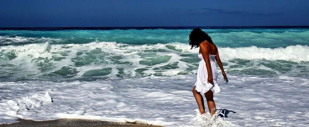 Eine junge Frau im weißen Kleid spielt mit den Füßen im Meer am Strand.