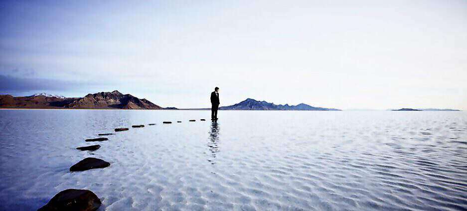 Ein Mann steht im Wasser auf einem Stein. Steine führen als Weg zum Wasser.