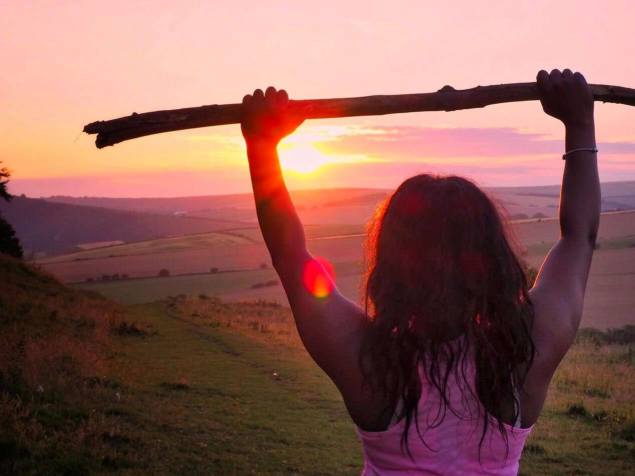 Eine junge Frau hält bei Sonnenuntergang einen Stock mit beiden Armen in die Luft.