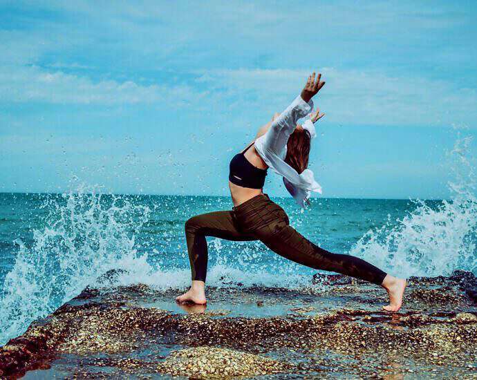 Eine junge Frau steht auf einem Felsen am Meer, während die Wellen auf sie einschlagen.