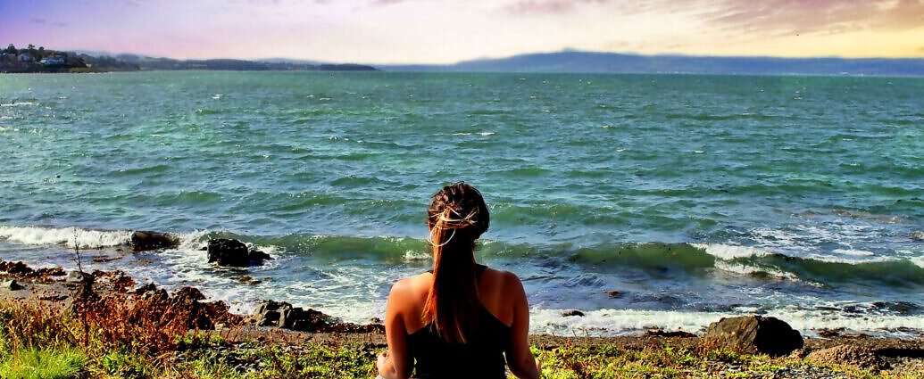 Eine junge Frau sitzt in Yoga-Kleidung auf einer Yogamatte am Meer und schaut entspannt auf das Wasser.