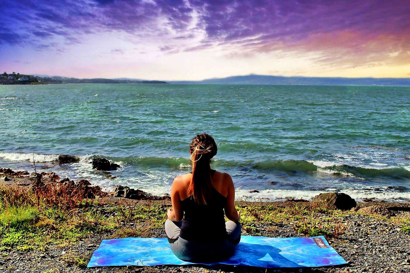 Eine junge Frau sitzt in Yoga-Kleidung auf einer Yogamatte am Meer und schaut entspannt auf das Wasser.