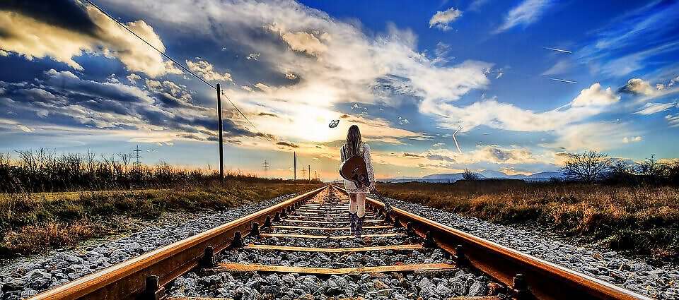 Eine junge Frau mit einer Gitarre auf dem Rücken geht ihren Weg auf einem Gleisweg.