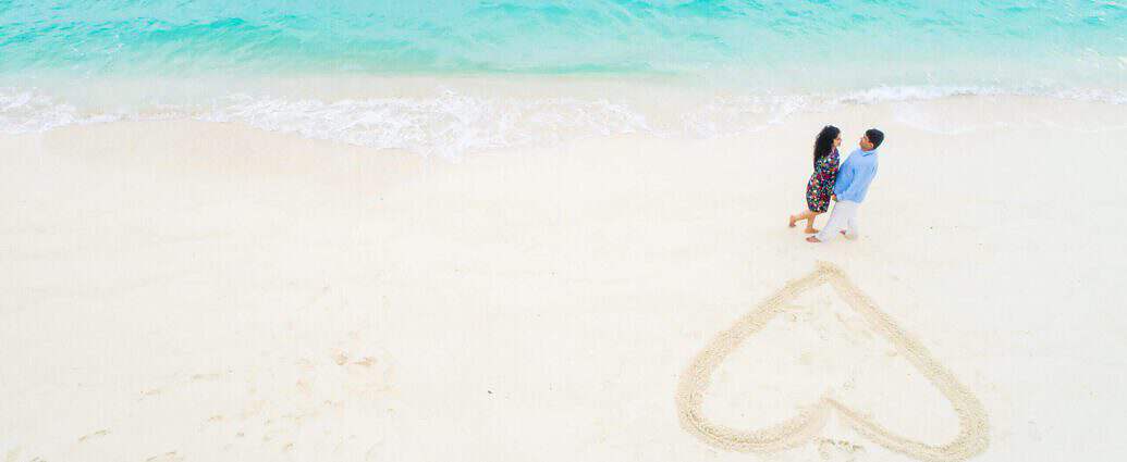 Ein junges, verheiratetes Paar schaut sich am Traumstrand verliebt an. Neben ihnen ist ein riesiges Herz aus Sand gemalt.