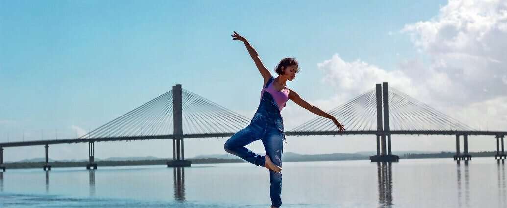 Eine junge Frau in Latzhose steht vor einer Hängebrücke und vollführt eine Ballettfigur.