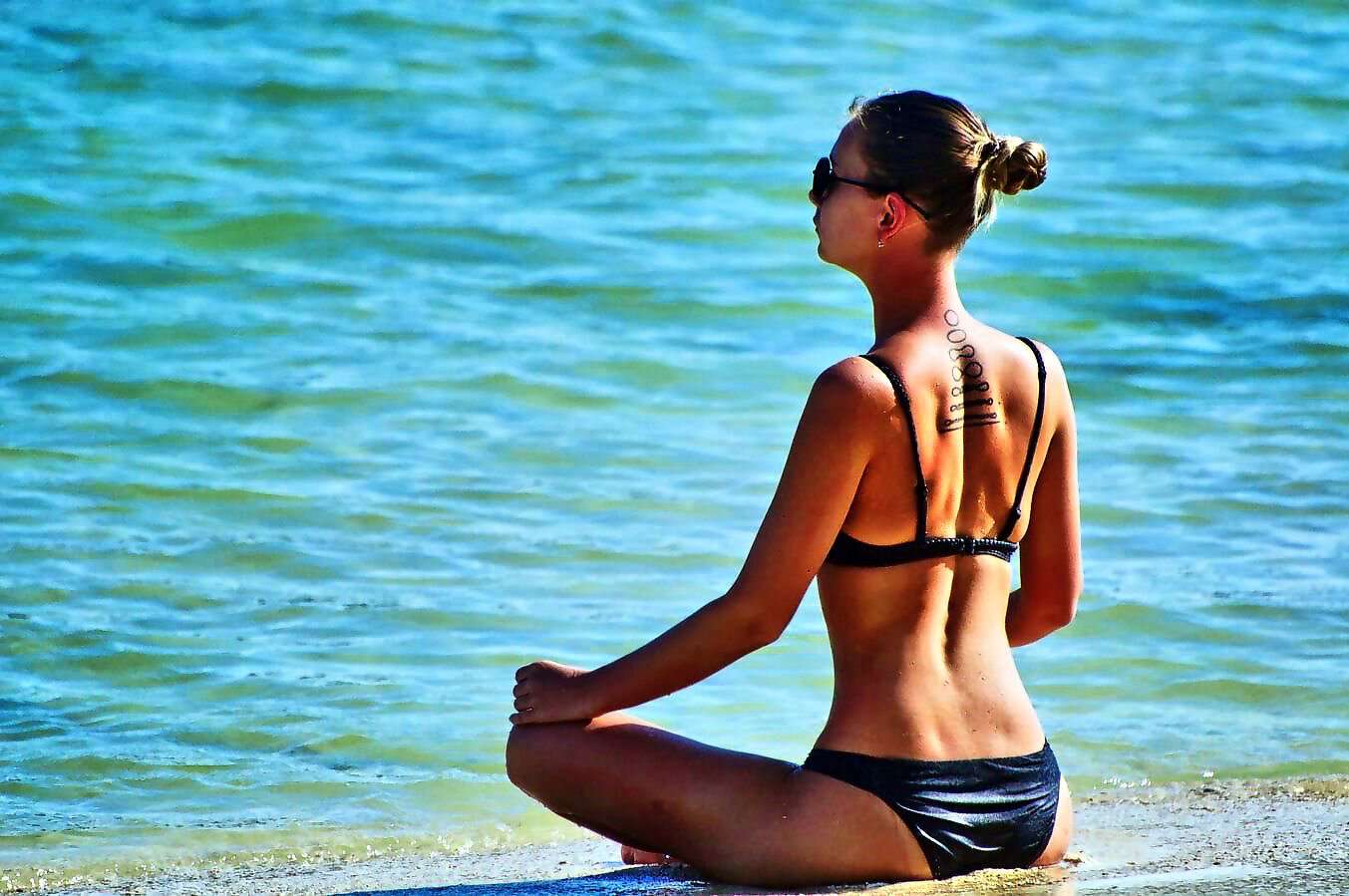 Eine junge Frau sitzt mit einem Bikini bekleidet am Strand und schaut entspannt auf das Meer.