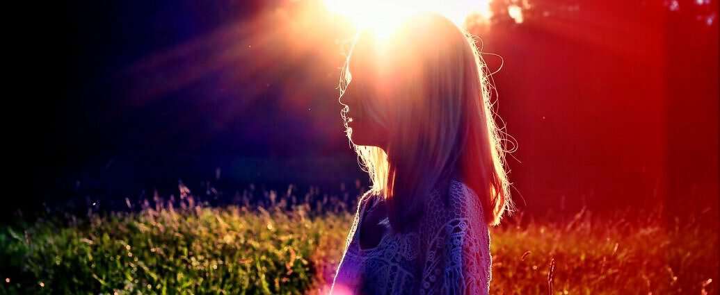 Zu sehen ist die Silhouette einer jungen Frau, die entspannt am Rande eines Waldes in der Abendsonne steht.