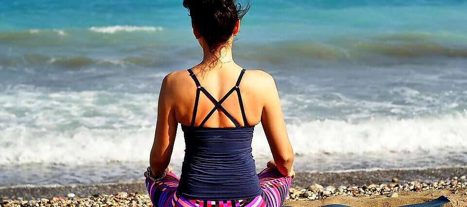 Eine junge Frau sitzt entspannt auf einer Yogamatte am Strand und schaut auf das Meer vor ihr.