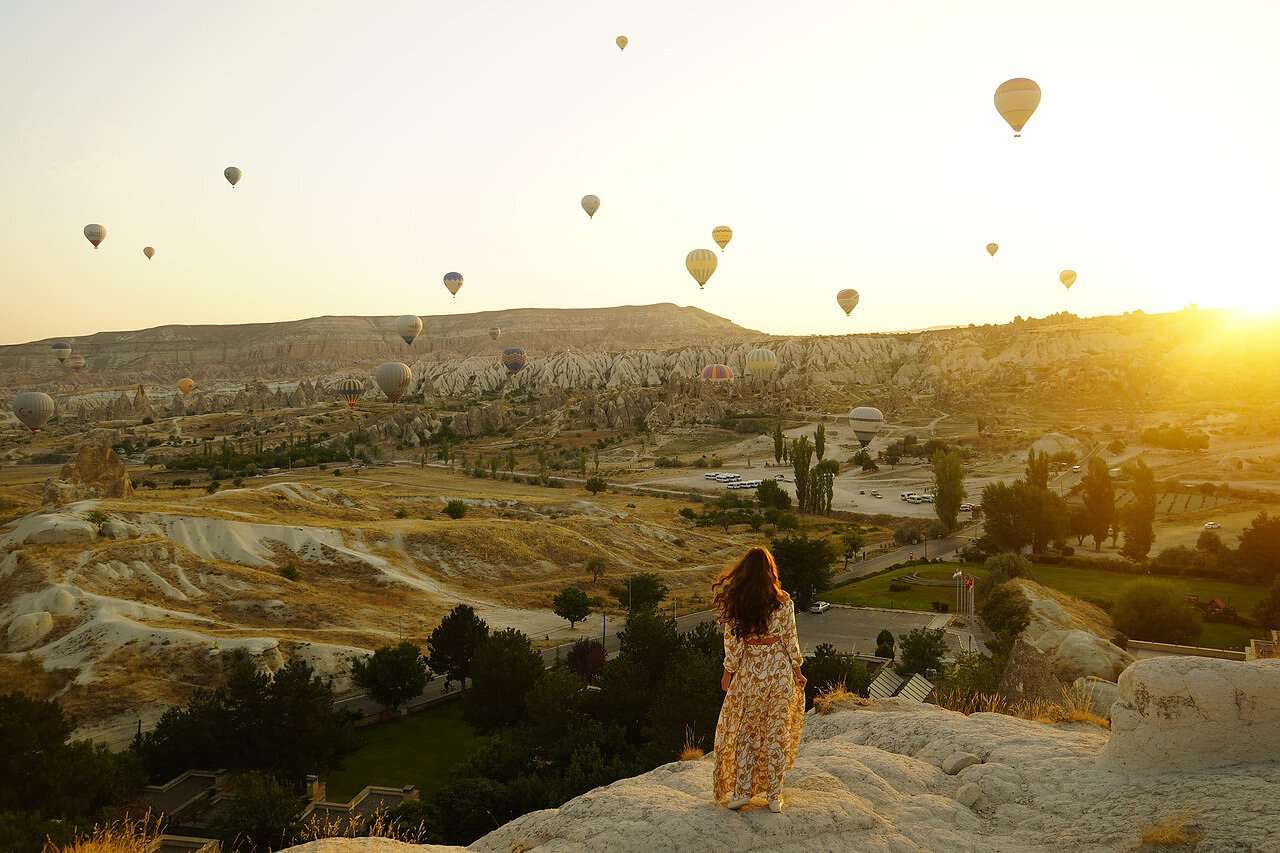 Eine Frau im Kleid steht auf einem Berghang und schaut auf Heißluftballons.
