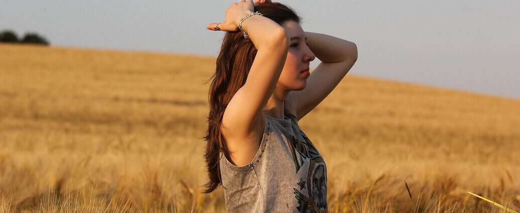 Eine junge Frau steht in einem Weizenfeld und verstrengt die Arme, um fröhlich durchzuatmen.