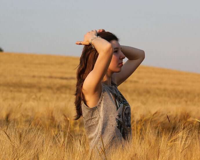 Eine junge Frau steht in einem Weizenfeld und verstrengt die Arme, um fröhlich durchzuatmen.