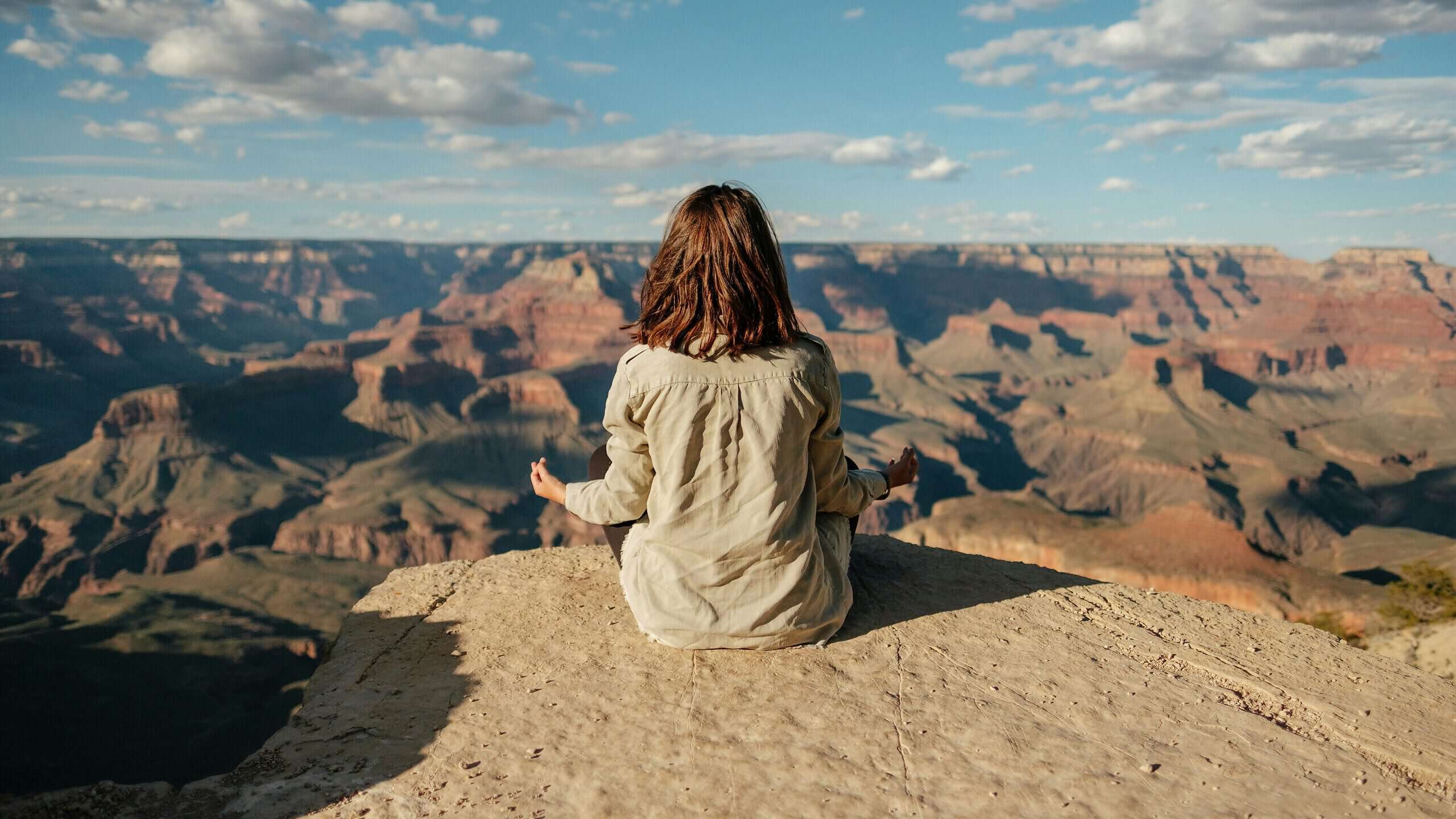 Eine junge Frau sitzt auf einem Felsvorsprung und meditiert. Vor ihr erstrecken sich die Berge und ein Canyon.