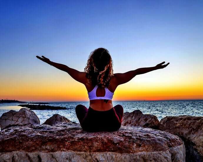 Eine junge Frau sitzt vor dem Meer in der Abendsonne und streckt die Arme aus.