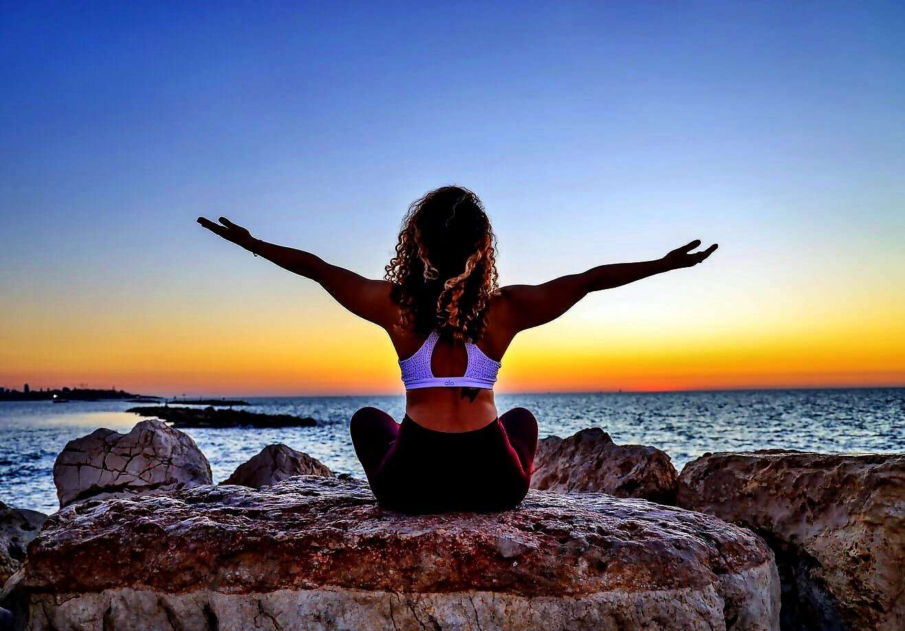 Eine junge Frau sitzt vor dem Meer in der Abendsonne und streckt die Arme aus.