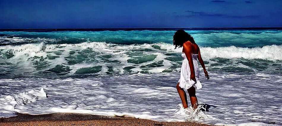 Eine junge Frau in einem weißen Kleid steht am Strand und hat die Füße im Meer.