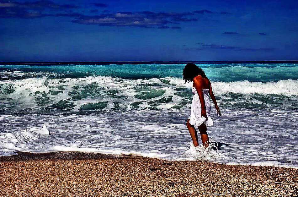 Eine junge Frau in einem weißen Kleid steht am Strand und hat die Füße im Meer.