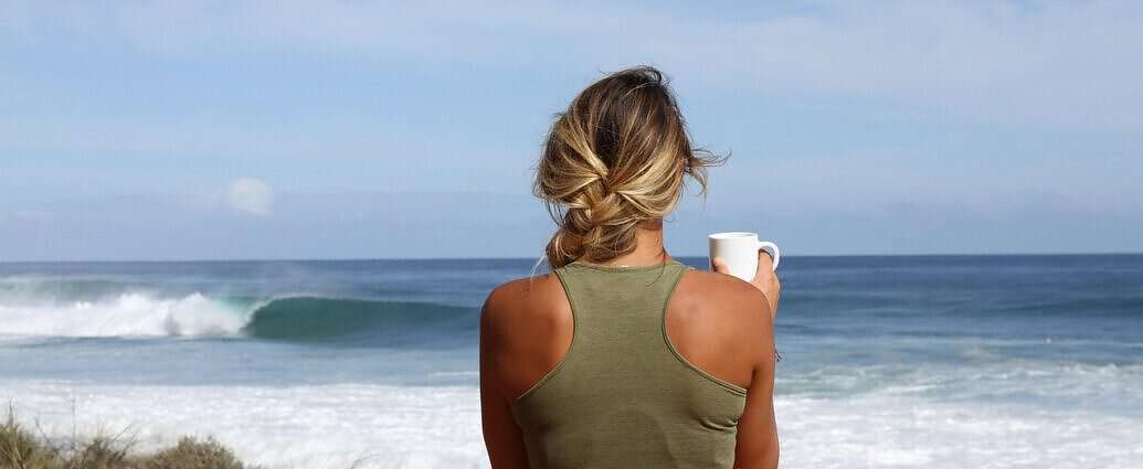 Eine junge Frau steht mit einer Tasse Tee am Strand und schaut in die Ferne.