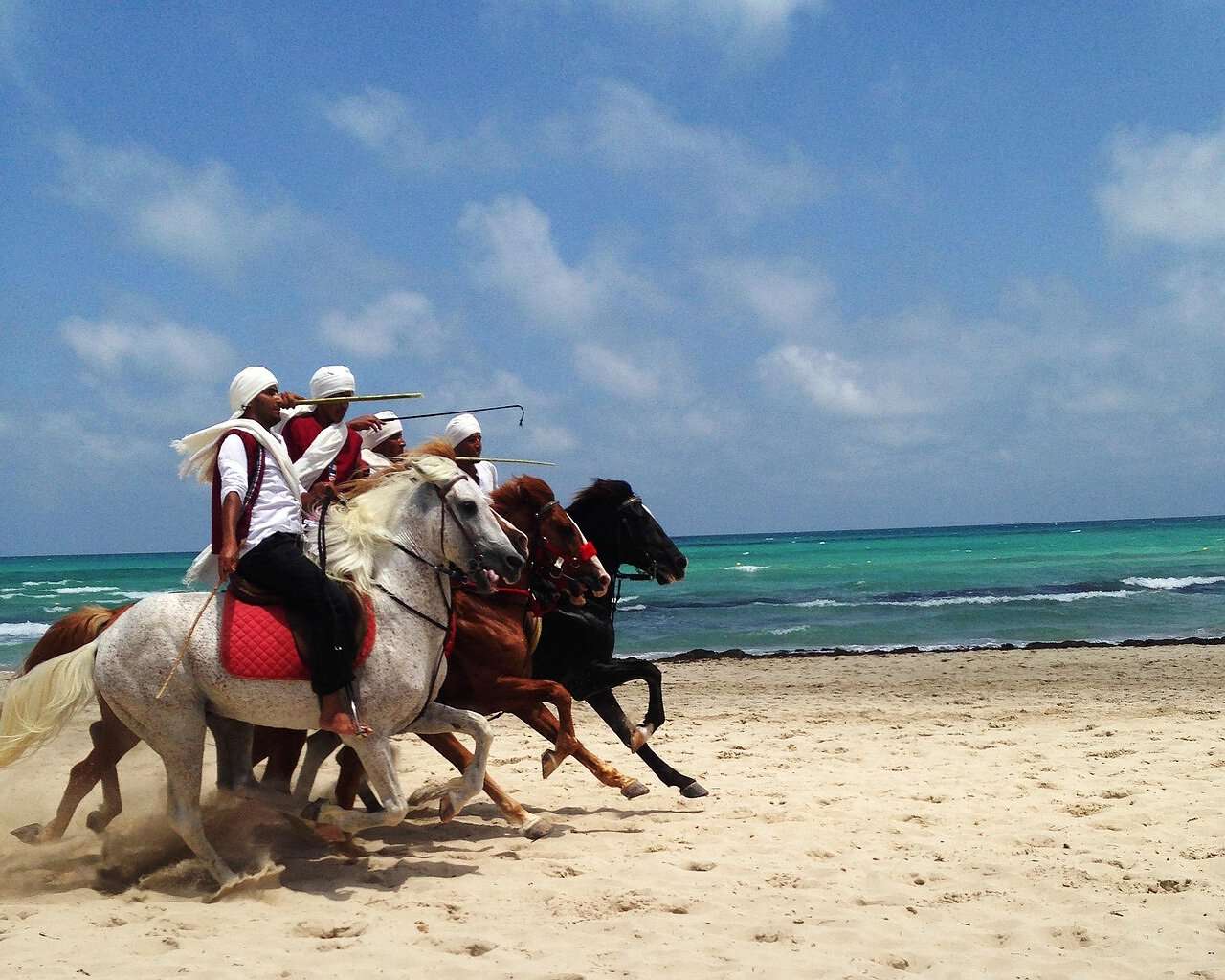 Ein Bild von tunesischen Einheimischen, die mit Pferden am Strand reiten.