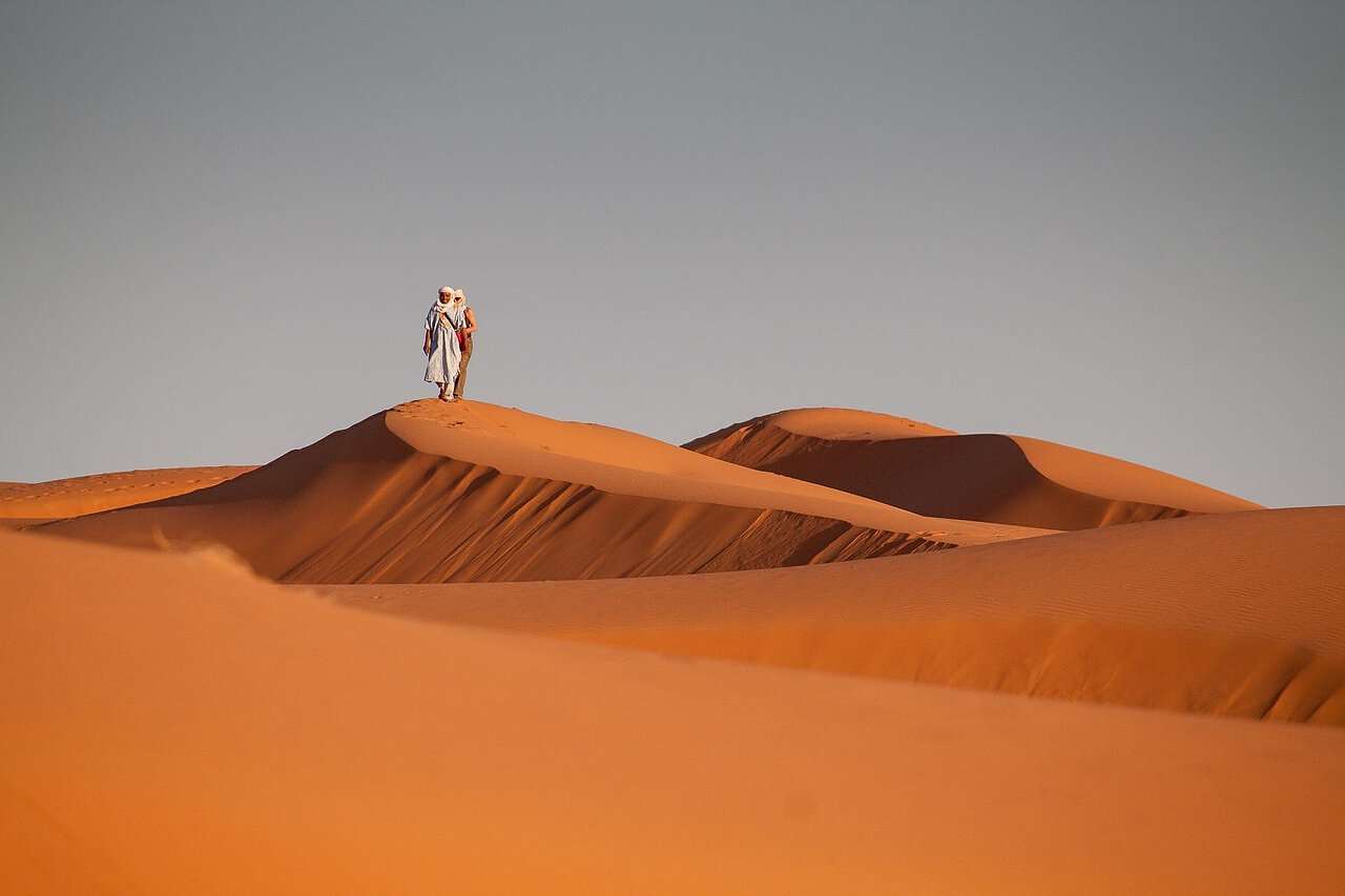 Ein Bild von zwei Beduinen, die in der marokkanischen Wüste auf einer Düne stehen.