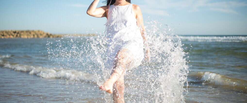 Eine Frau, die lachend am Ufer des Meeres steht und mit den Füßen im Wasser planscht.