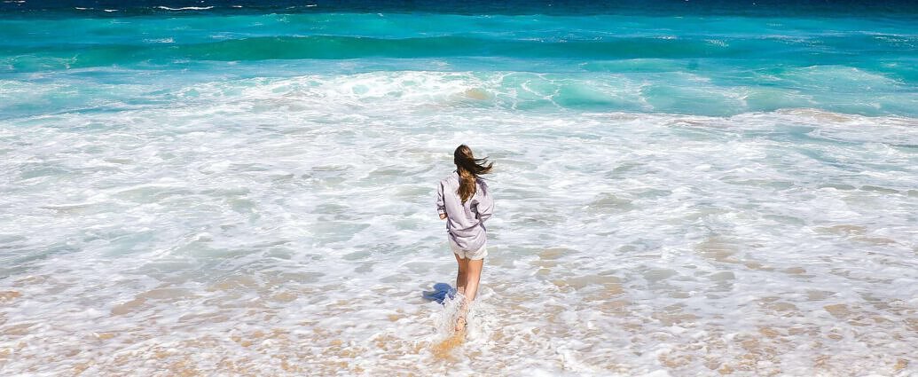 Ein Bild von einer jungen Frau, die am Strand im Wasser steht, mit einem weißen Strandkleid.