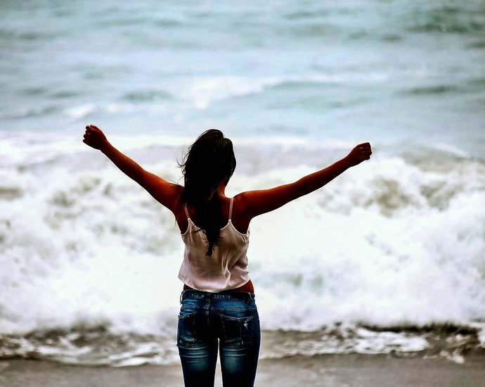Eine junge Frau hebt die Arme hoch, vor ihr ist das Meer.